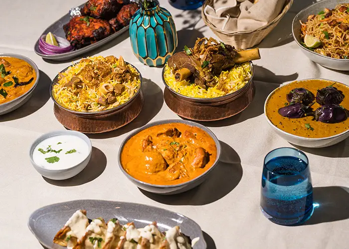 A laid table with mutton shank biryani, curries, tandoori chicken and a blue glass of water